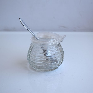 Peut inclure: Un pot à miel en verre transparent avec un corps arrondi et nervuré et un couvercle en verre assorti. Une petite cuillère en métal argenté avec un manche décoratif repose à l'intérieur du pot, dépassant de l'ouverture supérieure.