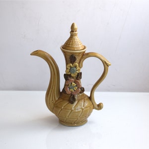 以下が含まれることがあります： 花柄の蓋が付いた茶色の陶器製のティーポット。ティーポットは織り模様と湾曲した取っ手のある注ぎ口があります。