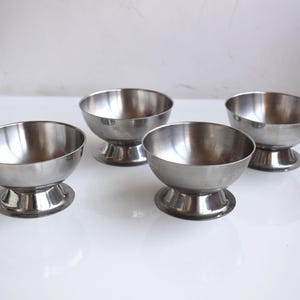 Op de afbeelding: Vier roestvrijstalen dessertkommen met een klassiek ontwerp. Elke kom heeft een afgeronde, gepolijste binnenkant en staat op een korte voet. De kommen zijn gerangschikt op een wit oppervlak.