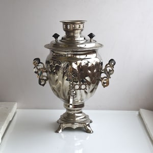 May include: A silver samovar with ornate details and a black knob on the lid. The samovar has a spout and two handles.