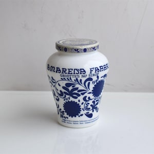 Può includere: Un barattolo di ceramica bianca con un motivo floreale blu e bianco. Il barattolo ha un coperchio e dice "Amarena Fabbri Griottes au Sirop" sul davanti.