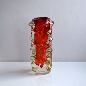 Könnte beinhalten: Eine rote und klare Glasvase mit einem strukturierten, wellenförmigen Design. Die Vase ist etwa 15 cm hoch.