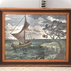 May include: A framed tapestry showing a sailboat on the water near a shoreline with a tree. The artwork features a brown sailboat with a brown sail, set against a cloudy sky. The frame is made of wood.