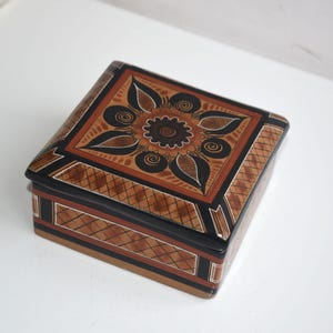 May include: Square wooden box with a floral design on the lid. The box features a brown and black colour scheme with intricate patterns. The lid has a central flower motif surrounded by leaf-like shapes. The sides of the box have a geometric pattern.