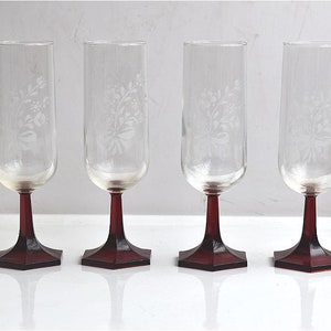 Puede incluir: Cuatro copas de vino de vidrio transparente con diseños florales grabados y tallos y bases de color burdeos.