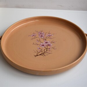 May include: A round, peach-colored serving tray with two small handles. The tray features a floral design of pink blossoms and brown branches in the center. The tray is likely made of ceramic or porcelain.