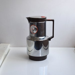 Puede incluir: Una jarra térmica de plata y marrón con un asa negra y un botón rojo en la tapa. La jarra tiene una pegatina con un diseño gráfico en el lateral.