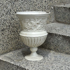 May include: A white ceramic planter with a decorative floral design. The planter has a wide base and a narrow top, and it is sitting on a set of concrete steps.