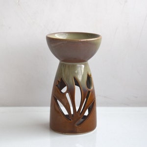Può includere: Un bruciatore per oli essenziali in ceramica marrone e verde con un design floreale. Il bruciatore ha una ciotola larga e poco profonda in cima e una base con fiori ritagliati.