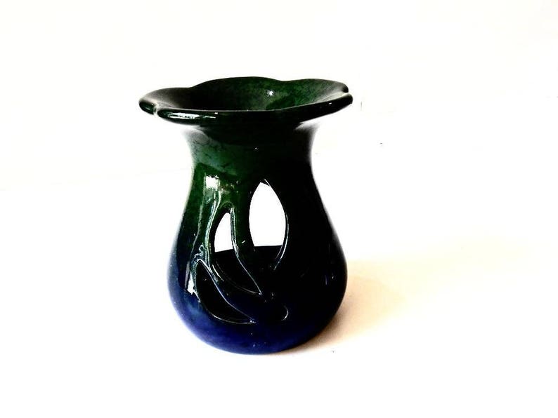 May include: A green and blue ceramic oil burner with a decorative cutout design. The burner has a wide, flat top and a rounded base.