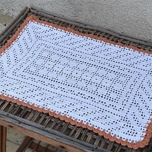 May include: White crocheted table runner with a peach-colored border. The runner is laying on a woven table top.