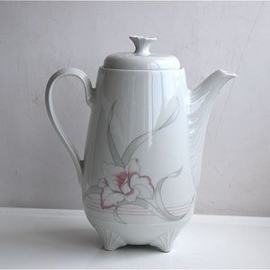 Può includere: Caffettiera in ceramica bianca con un motivo floreale. La caffettiera ha un coperchio e un beccuccio. Il motivo floreale presenta un fiore rosa con foglie verdi.