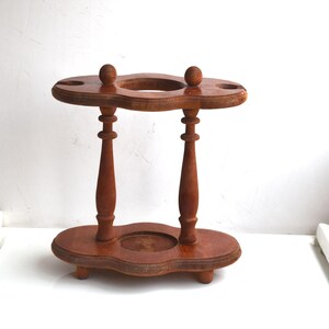 Può includere: Un portapipe vintage in legno con finitura marrone scuro. Il supporto presenta un design a due livelli con una base circolare e una parte superiore con due fori e due piccoli accenti a forma di sfera. Il supporto è sostenuto da due colonne tornite.