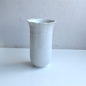 May include: A white porcelain vase featuring a vertical ribbed texture and a scalloped rim. The cylindrical body has a smooth finish with subtle embossed wave patterns near the top opening, creating a clean and uniform appearance throughout the piece.