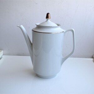 Puede incluir: Una cafetera de porcelana blanca con un cuerpo cilíndrico alto y detalles acanalados verticales. La cafetera incluye una tapa a juego con un remate metálico en tono dorado, un asa curva y un pico largo, acentuado por un fino borde dorado.
