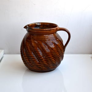 May include: A round, brown glazed ceramic pitcher featuring a distinct spiral, ribbed texture across its body. The vessel has a wide, open mouth with a small pouring spout and a single curved handle attached to the side, showcasing a glossy finish.
