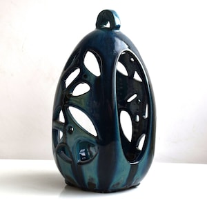 May include: Teal blue ceramic candle holder with a unique cutout design. The holder is shaped like an egg and has a hole in the center for a candle.