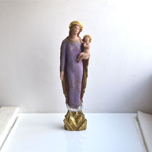 Peut inclure: Une statue peinte en violet et or de la Vierge Marie tenant un bébé Jésus. La statue est posée sur une base dorée.