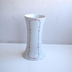 Könnte beinhalten: Eine weiße Keramikvase mit ausgestelltem Fuß und gewelltem Rand. Die Vase ist mit einem rosa Blumenmuster verziert, das vertikal an den Seiten verläuft. Die Vase ist etwa 20 cm hoch.