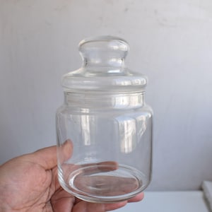 Puede incluir: Un tarro de vidrio transparente con tapa. El tarro es cilíndrico con una base redondeada y una parte superior ligeramente más ancha. La tapa es de vidrio transparente a juego con un pequeño pomo. El tarro está sostenido por una mano.