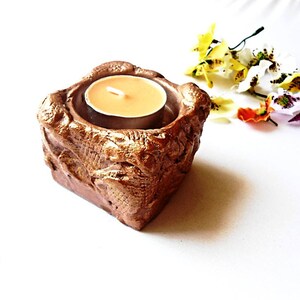 May include: A square, textured, bronze-coloured candle holder with a lit tea light candle inside. The candle holder has a rough, organic appearance. Decorative flowers in yellow, white, orange, and purple are arranged nearby.