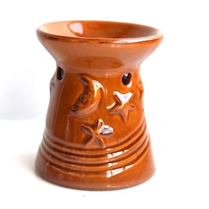 Pode incluir: Um queimador de óleo de cerâmica marrom com um design de lua e estrelas. O queimador tem um pequeno poço na parte superior para conter óleo e um pequeno furo na lateral para inserir uma vela de chá.