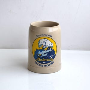 Puede incluir: Una jarra de cerveza de cerámica beige con una ilustración azul y amarilla de una mujer con un sombrero blanco y el texto "Jahrskrug 1988", "Deutsches Bier", "Hacker-Pschorr" y "1928".