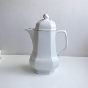 Puede incluir: Cafetera de porcelana blanca con tapa y asa. La cafetera tiene un cuerpo redondeado y una base cuadrada.