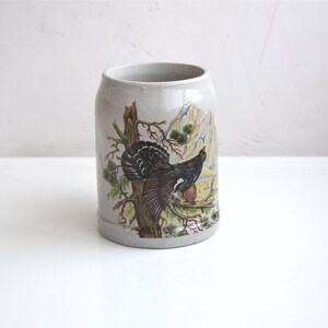 May include: A grey ceramic beer stein featuring a detailed illustration of a bird on a tree branch, with mountains in the background. The stein is cylindrical with a wide opening.