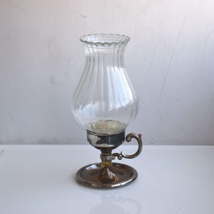 Può includere: Un portacandele a lanterna in vetro trasparente con base e manico in metallo color argento. Il vetro ha delle scanalature verticali e un bordo smerlato. La base è rotonda con un piccolo manico laterale.