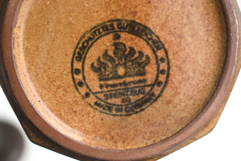 Może przedstawiać: Zbliżenie na brązowy ceramiczny garnek z czarną pieczęcią, na kt&oacute;rej widnieje napis "Geschuetztes Warenzeichen" i "Steingut Made in Germany".