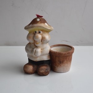May include: A whimsical ceramic planter shaped like a mushroom-headed gnome. The gnome wears a white shirt, black pants, and brown shoes. A small brown pot sits beside the gnome. The planter is painted in earthy tones with a red accent.