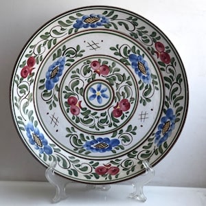 Może przedstawiać: Biały ceramiczny talerz z dekoracyjnym wzorem niebieskich i czerwonych kwiatów oraz zielonych liści. Talerz stoi na białym stojaku.