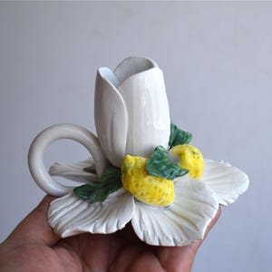 Puede incluir: Candelabro de cerámica blanca con forma de flor y centro en forma de tulipán. El portavelas presenta limones amarillos y hojas verdes. El portavelas tiene un asa circular y está diseñado para una sola vela.