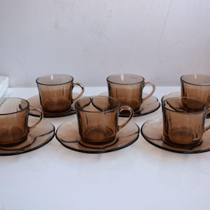 May include: Six amber-colored glass teacups and saucers. Each cup has a handle and sits on a matching saucer. The cups and saucers are arranged in two rows of three, showcasing a vintage tea set.