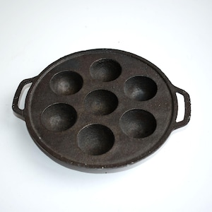 May include: A round, dark grey cast iron pan featuring seven circular indentations for cooking. The pan has two integrated side handles and a textured, matte surface finish. The design includes one central circular mould surrounded by six others.