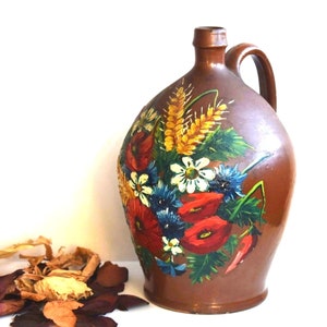 May include: A brown ceramic jug with a floral design painted on the front. The flowers are red, white, blue, and yellow. The jug has a handle and a wide mouth.