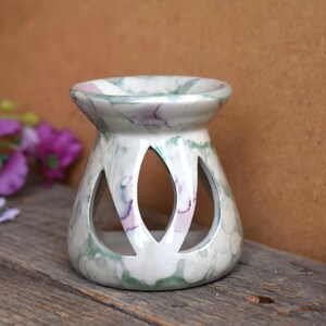Può includere: Un bruciatore per oli in ceramica bianca con un design marmoreo in tonalità di verde e rosa. Il bruciatore ha una parte superiore rotonda e una base larga con tre aperture tagliate.