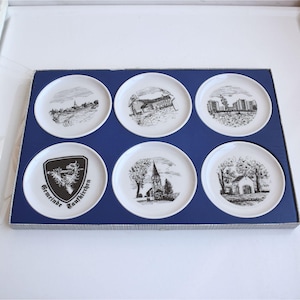 May include: A set of six white ceramic coasters with black and white illustrations of buildings and landscapes. The coasters are arranged in a blue box, perfect for home decor or a unique gift.