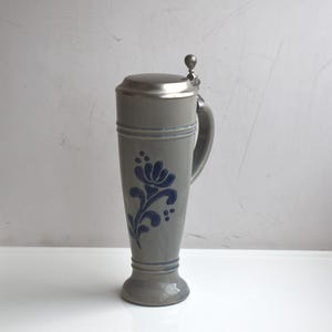 Peut inclure: Chope à bière en céramique grise avec un motif floral bleu et un couvercle à charnière en métal. La chope a une anse et mesure environ 25 cm de haut.