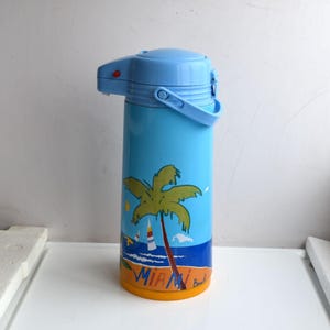 Könnte beinhalten: Eine hellblaue Isolierflasche mit Palmen- und Strandszene-Design. Der Behälter hat einen blauen Griff und Deckel, und die Worte "Miami Beach" sind auf der Unterseite aufgedruckt.