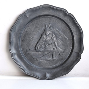 Op de afbeelding: Een tinnen wandbord met een reliëf van een paardenkop. Het paard draagt een hoofdstel en kijkt naar links. Het bord is donkergrijs en heeft een gegolfde rand.