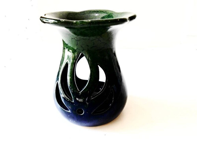 May include: A green and blue ceramic oil burner with a decorative cutout design. The burner has a wide, flat top and a round base.