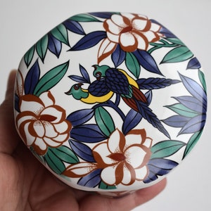 May include: A white ceramic lid with a floral design featuring blue, green, yellow, and red birds and flowers.