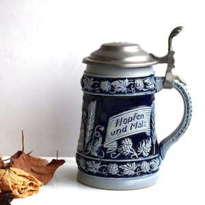 May include: A blue and white ceramic beer stein with a pewter lid and handle. The stein is decorated with a floral design and the text "Hopfen und Malz".