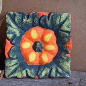 May include: A ceramic tile with a hand-painted design of a flower with orange, yellow, and black petals. The tile has a textured surface and is a decorative accent.