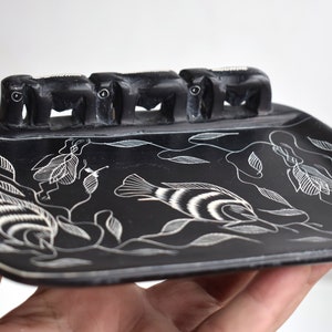 May include: Black soap dish with a carved design of fish and leaves. The dish has four small elephant figures on the edge.