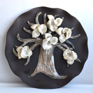 May include: A ceramic wall hanging featuring a stylised tree with white flowers and silver branches on a dark brown background.