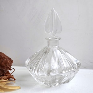 May include: A clear glass bottle with a faceted design and a decorative stopper. The bottle has a square base and a rounded body.