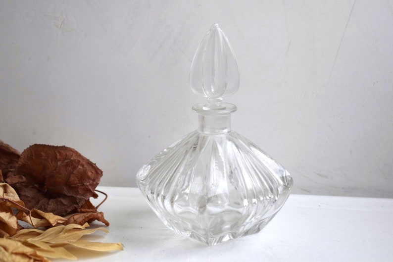 May include: A clear glass bottle with a faceted design and a decorative stopper. The bottle has a square base and a rounded body.